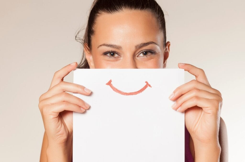 Junge fröhliche Frau hält ein Blatt Papier mit einem Lächeln darauf vor ihr halbes Gesicht.